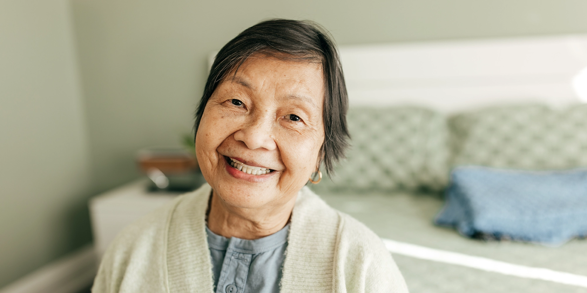 An elderly woman smiles tenderly. She's sitting on the edge of her bed in her own home.