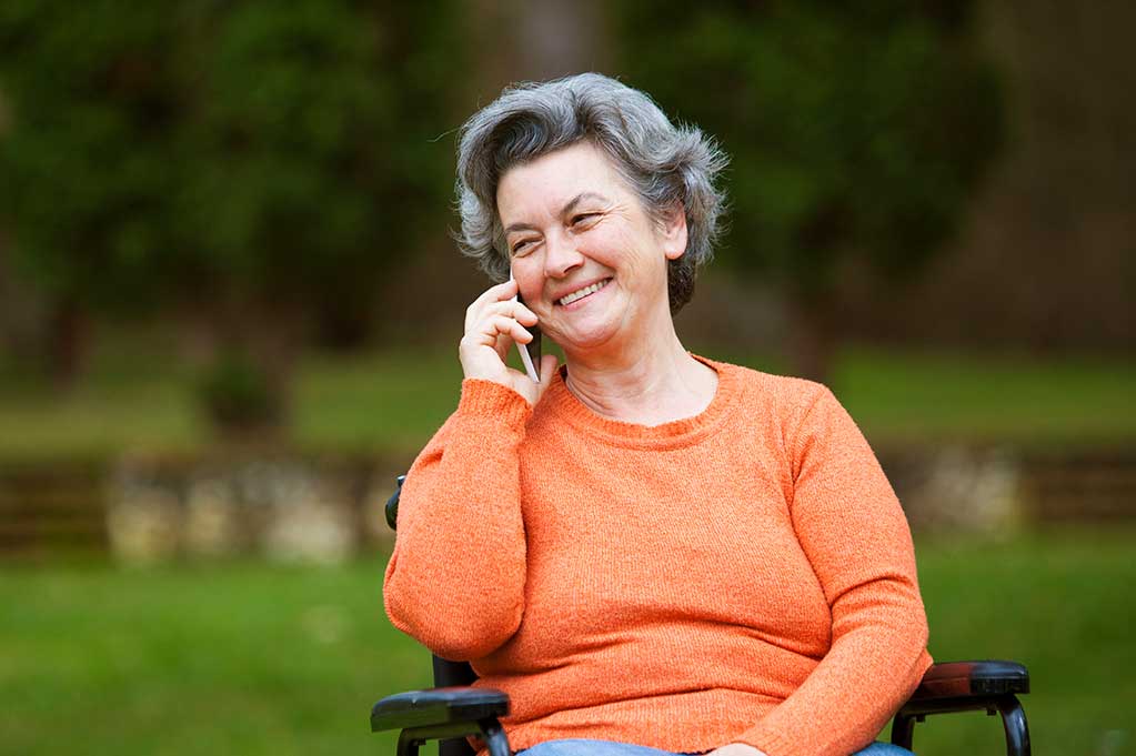 An older woman happily calls someone on the phone. She's sitting on a park bench.