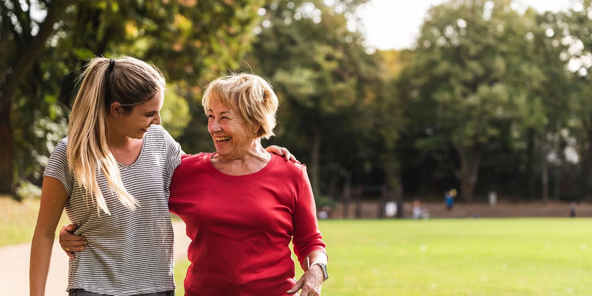 Two generations of women embrace in a park. It's sunny outside and they're in an open grassy area.