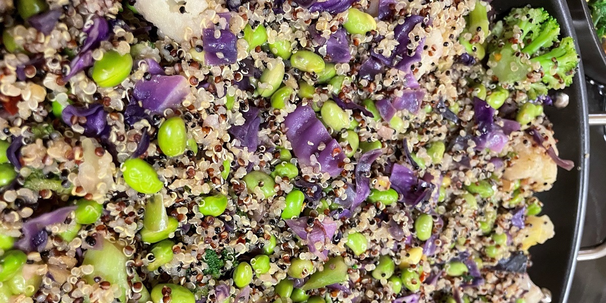 Colorful stir-fry of greens and purples made using edamame, broccoli florets, quinoa, and purple cabbage in a cast iron wok.